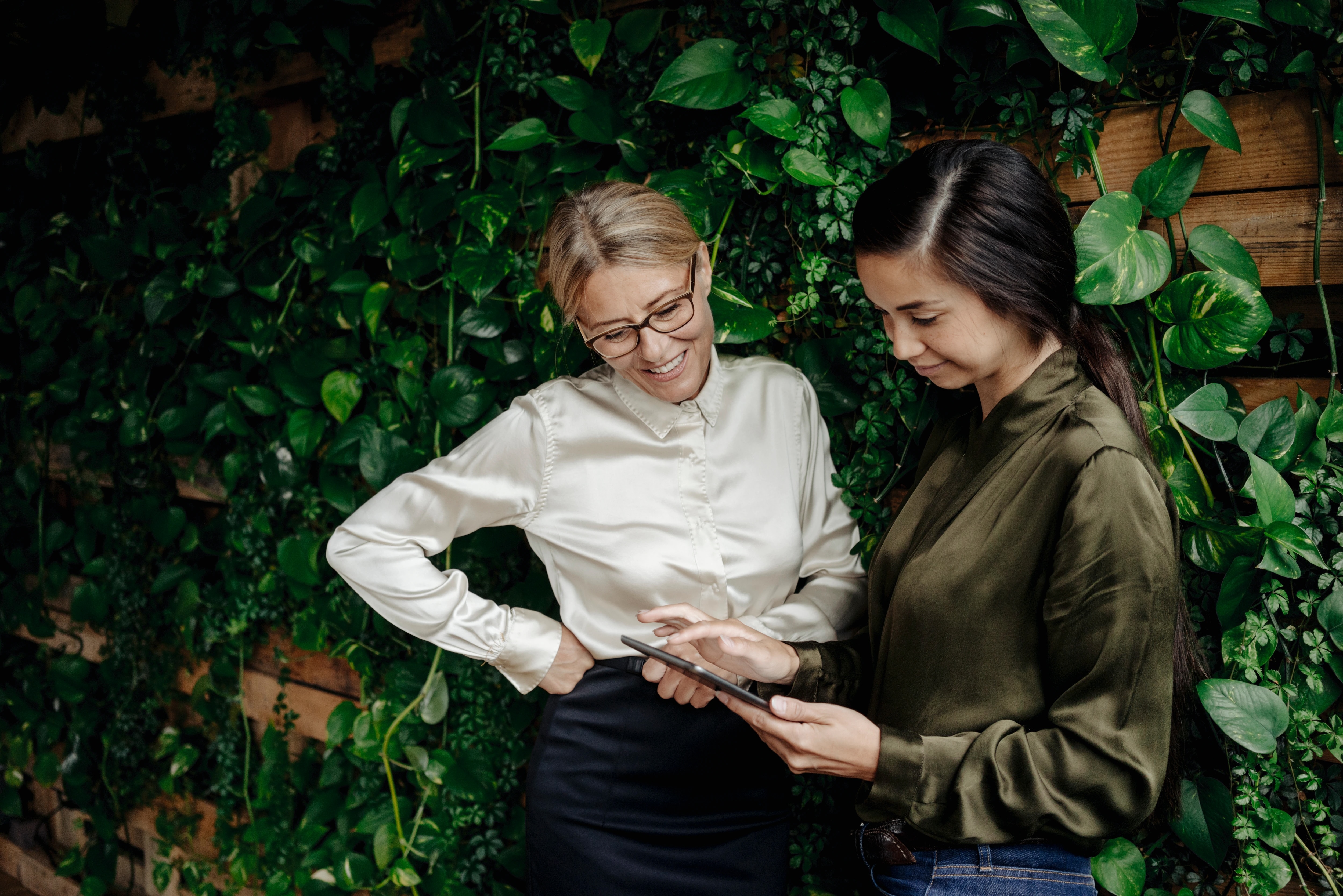 Two women looking at tablet with an ivy-covered fence behind them